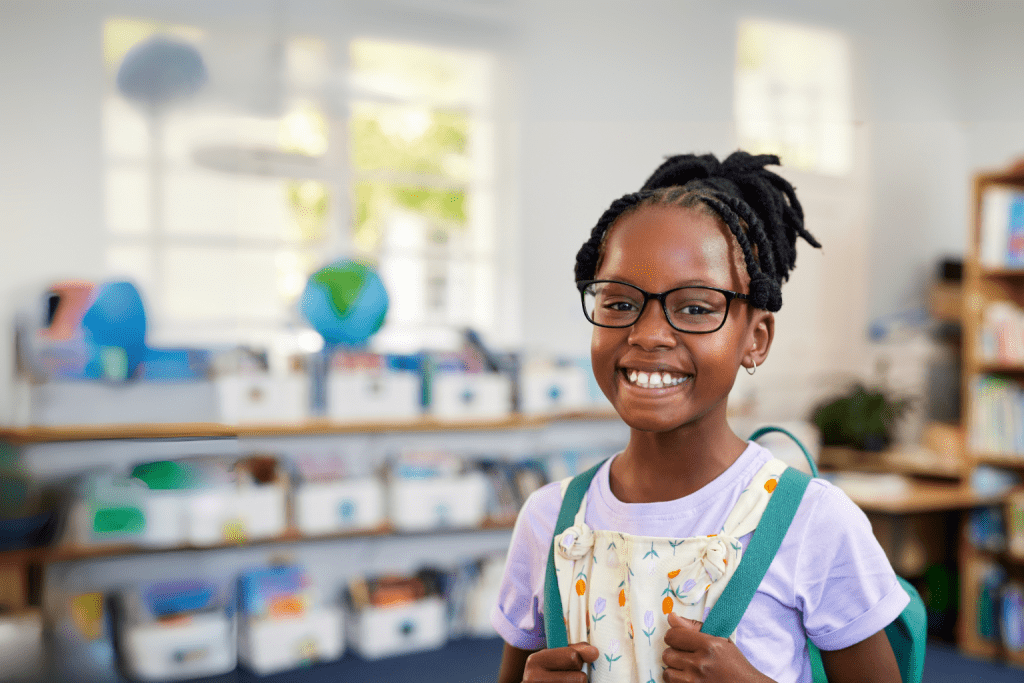 Smiling elementary school student wearing glasses and a backpack, standing in a brightly lit classroom with books and educational materials in the background.